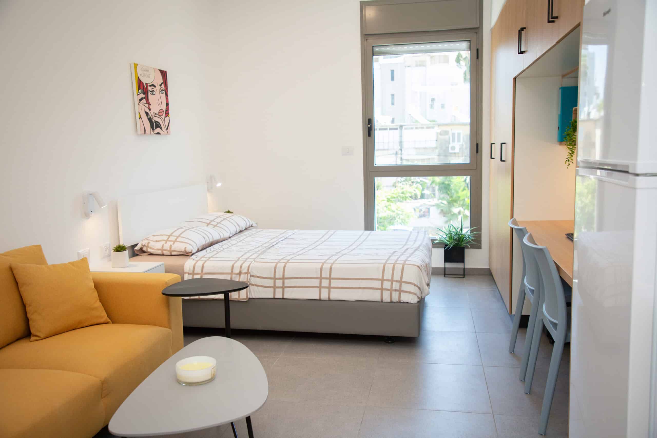 דירת סטודיו מוארת עם מיטה זוגית, ספה בצבע חרדל, שולחן קפה מינימליסטי, שולחן עבודה, חלון גדול ודלת ארון בצבע עץ בהיר.