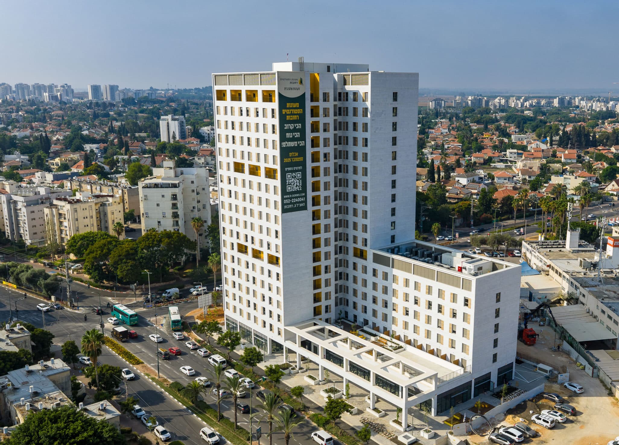 בניין רב קומות לבן עם חלונות רבות במרכז נוף עירוני עם כבישים ועצים, כאשר מבנים נמוכים יותר נראים ברקע.