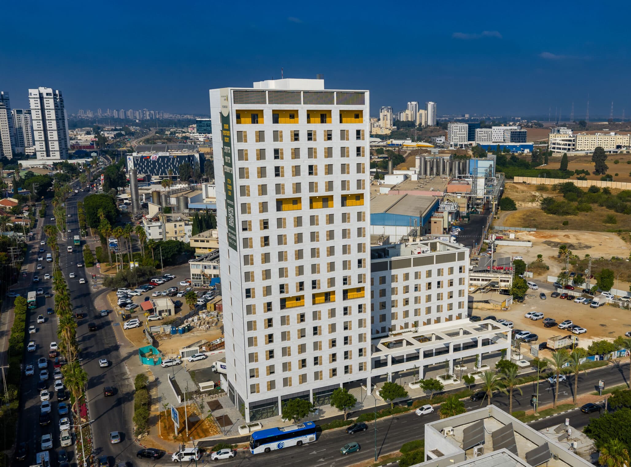 בניין רב קומות לבן עם חלונות צהובים, על רקע נוף עירוני ושטח פתוח, עם רכבים בכביש שלצידו.