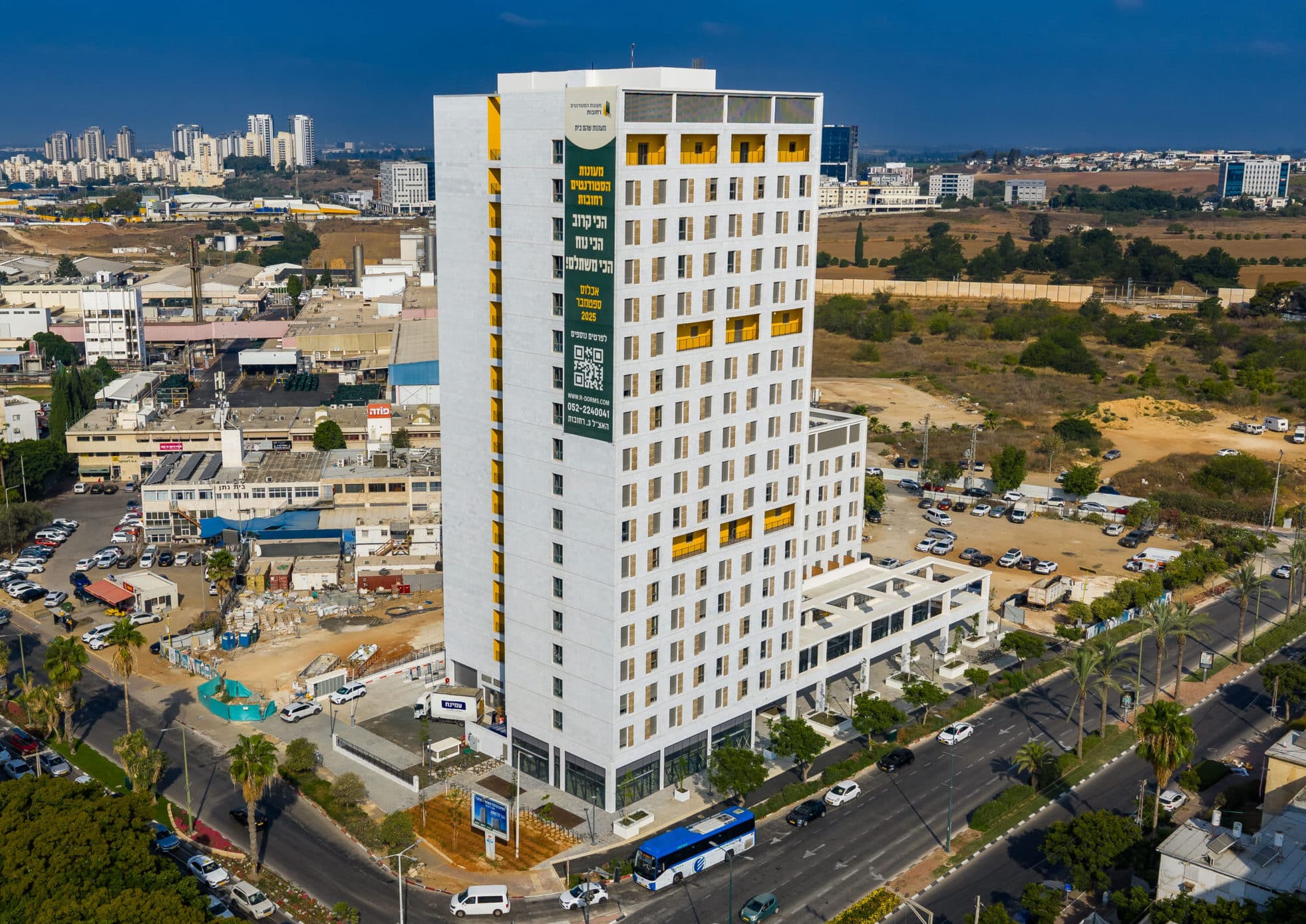 בניין רב קומות לבן עם חלונות צהובים, ממוקם בצומת רחובות, עם בניינים ומבנים מסחריים מסביב.