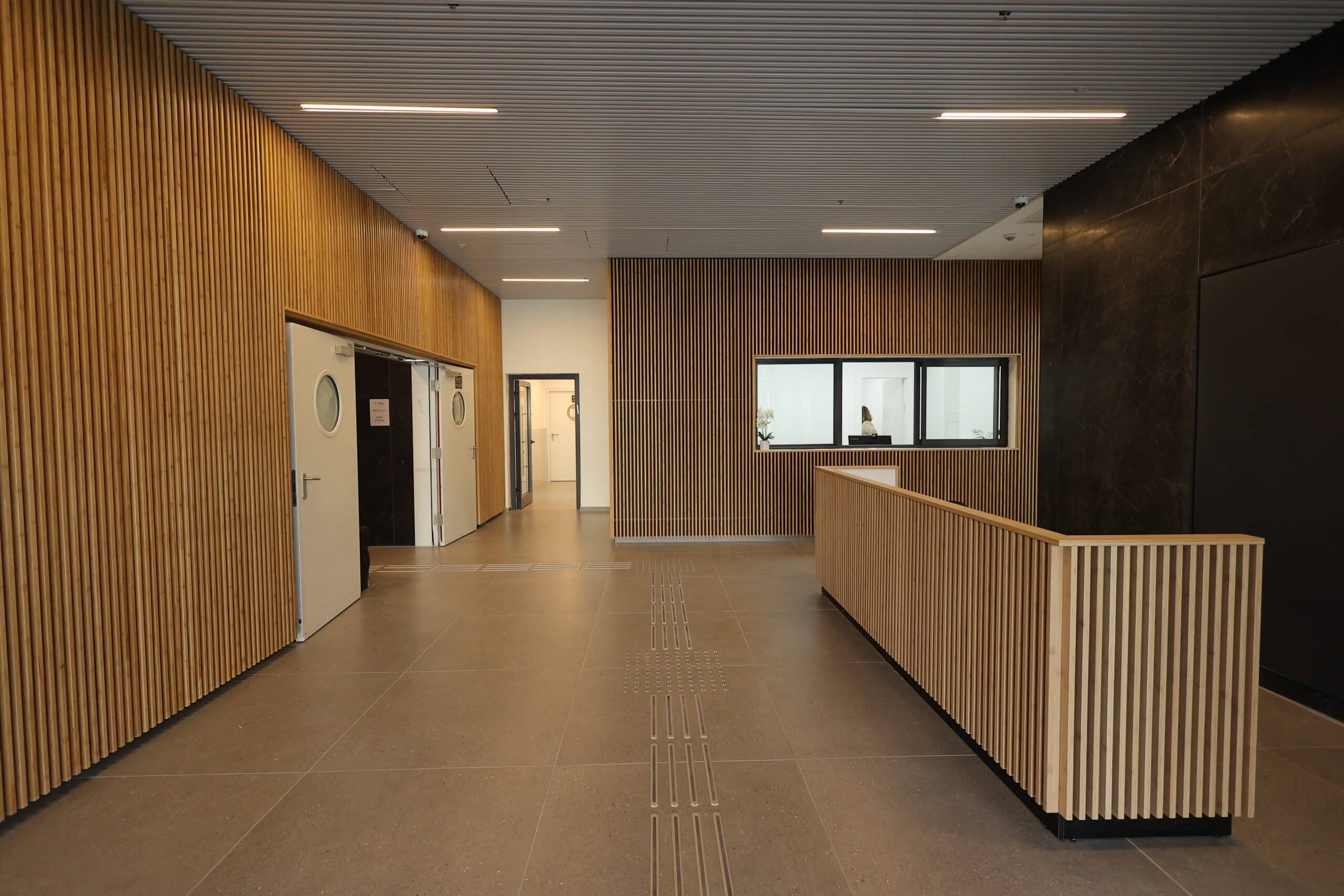 לובי מודרני עם קירות ותקרה בציפוי עץ, דלפק קבלה מעץ, דלתות לבנות עם חלונות עגולים ואדם יושב מאחורי דלפק זכוכית.