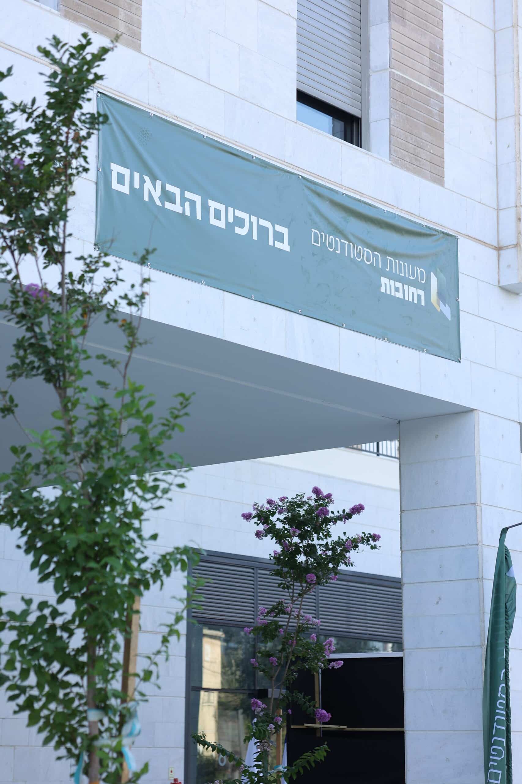 שלט בכניסה לבניין עם הכיתוב 'ברוכים הבאים' ומועדון הסטודנטים רחובות, עם צמחים פורחים לצידו.