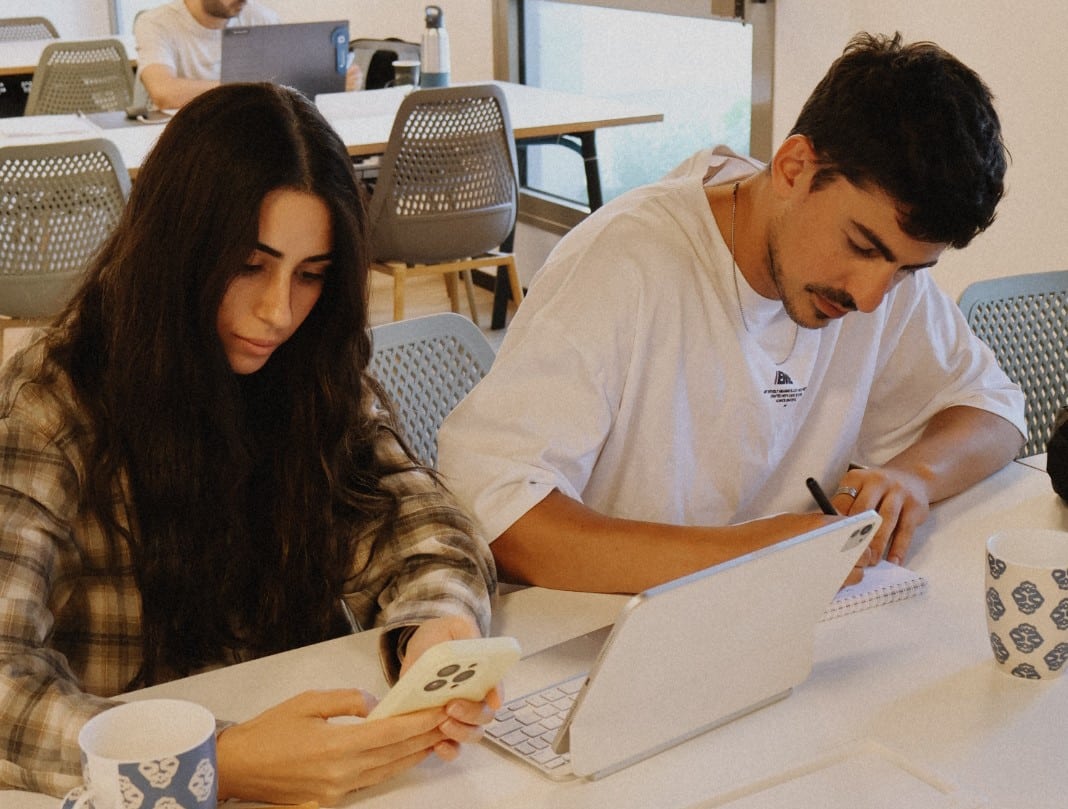 אישה עם שיער כהה וגבר עם חולצה לבנה יושבים ליד שולחן. האישה מחזיקה טלפון נייד והגבר כותב במחברת. מסביבם כוסות וגאדג'טים על השולחן.